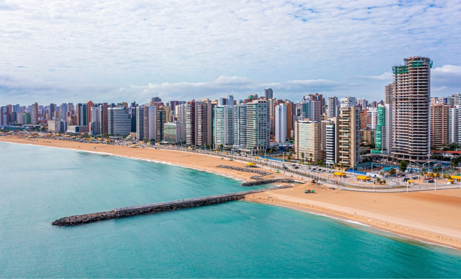 Fortaleza Orla de Fortaleza com edifícios altos à beira-mar, faixa de areia ampla, mar verde-azulado e píer de pedras sob céu parcialmente nublado.