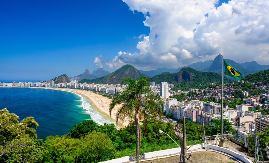 Rio de Janeiro Vista panorâmica da Praia de Copacabana, com mar azul, prédios à beira-mar, morros ao fundo e bandeira do Brasil ao lado direito sob céu com nuvens.
