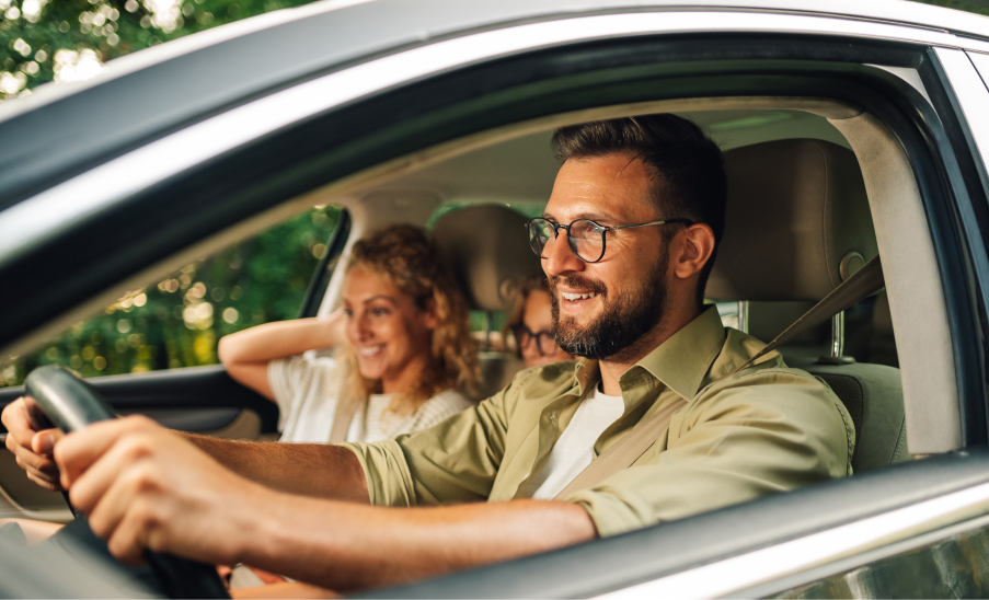 Autos Hombre al volante con acompañantes sonriendo, ventana con verde exterior.