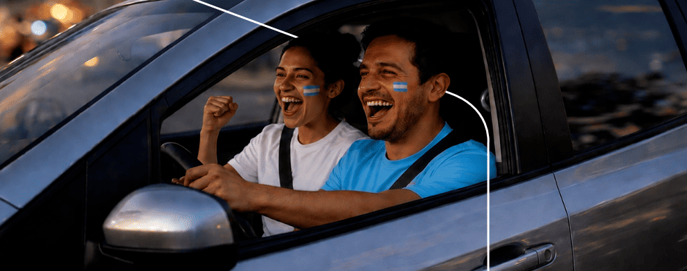 Autos Pareja con camisetas de Argentina riendo dentro de un auto al atardecer, vista lateral del conductor.