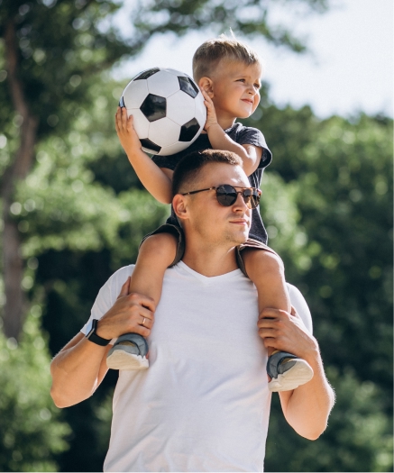 Seguros Adulto llevando a un niño en hombros con pelota de fútbol, día soleado.