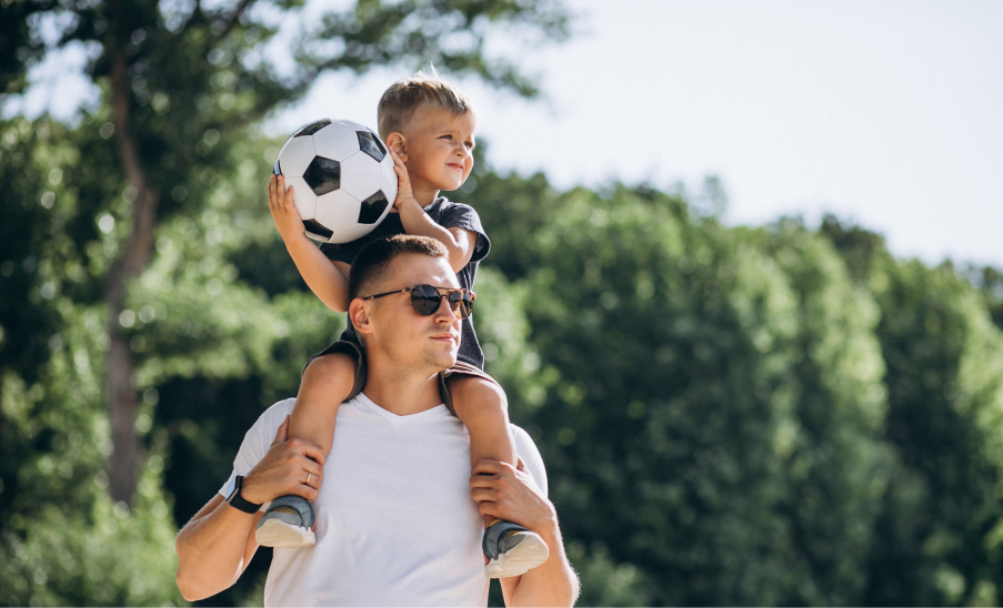 Seguros Adulto llevando a un niño en hombros con pelota de fútbol, día soleado.