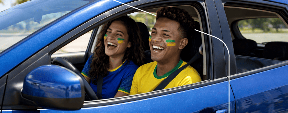 Casal com camisetas do Brasil rindo dentro de um carro ao pôr do sol, vista lateral do motorista