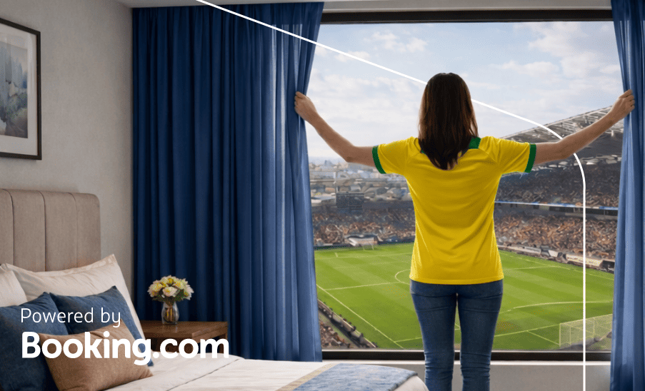 Mulher abre cortinas de um quarto de hotel com vista direta para um estádio; texto “Powered by Booking.com”.