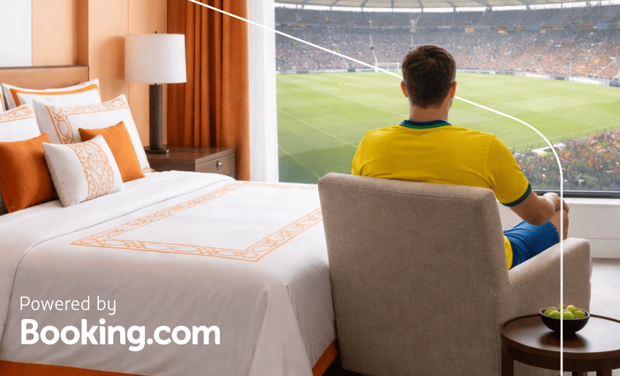 Homem sentado junto à janela de um quarto de hotel com vista para o gramado; texto “Powered by Booking.com”.