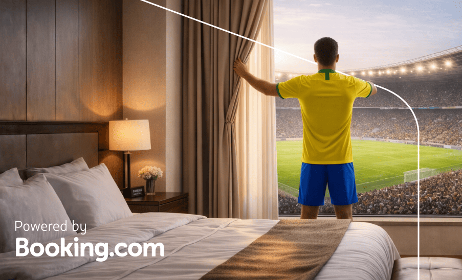 Homem com camiseta amarela de costas olhando o estádio da janela de um quarto de hotel; “Powered by Booking.com”