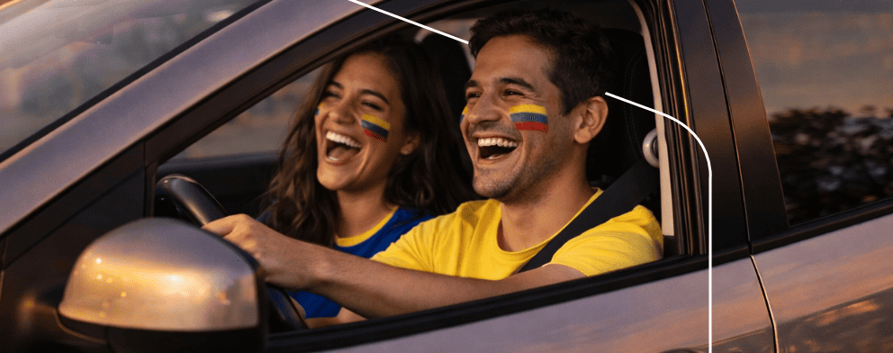 Pareja con camisetas de Colombia riendo dentro de un auto al atardecer, vista lateral del conductor.