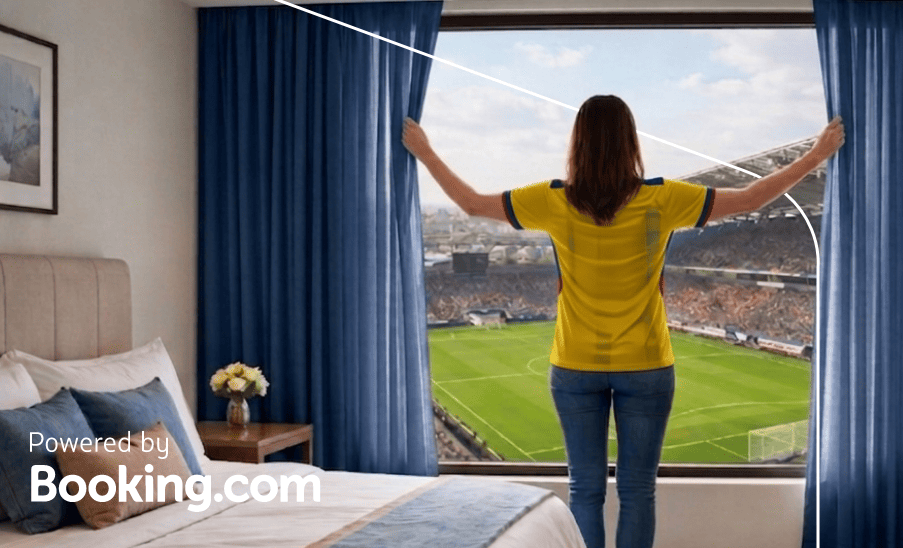 Kansas Mujer abre cortinas de una habitación de hotel con vista directa a un estadio; texto “Powered by Booking.com”.