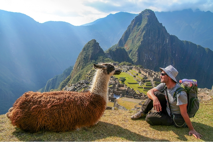What to pack - Andean Highlands Hiker in breathable trekking clothes and sun hat sitting with backpack next to a llama, overlooking Andean mountain ruins.