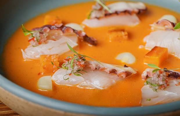 Cosme Peruvian seafood dish in an orange ají sauce with thin fish slices, octopus, and sweet potato in a blue bowl.