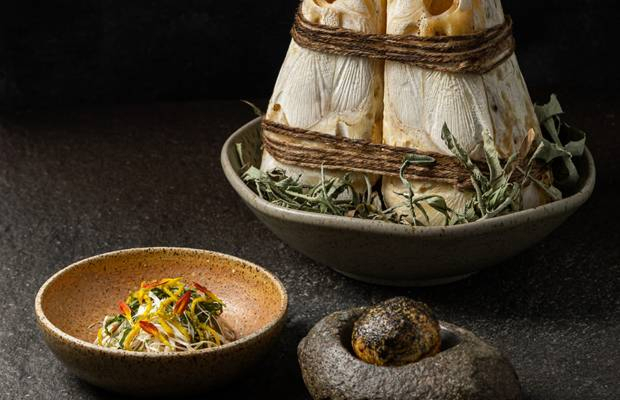 Mayta Modern Peruvian presentation with corn-husk bundles, a bowl of Andean noodles, and rustic stone on a dark table.